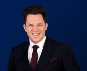 Rechtsanwalt Falco Schulz, Fachanwalt für Bau- und Architektenrecht sowie Erbrecht, in dunkelblauem Doppelreiher-Anzug mit roter Krawatte vor dunkelblauem Hintergrund.