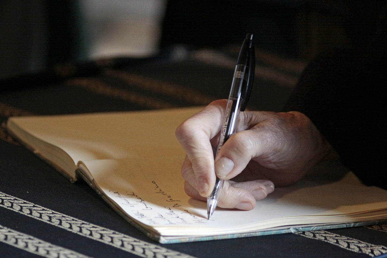 In dem Bild ist eine mit Kugelschreiber in ein Notizbuch schreibende Hand abgebildet.