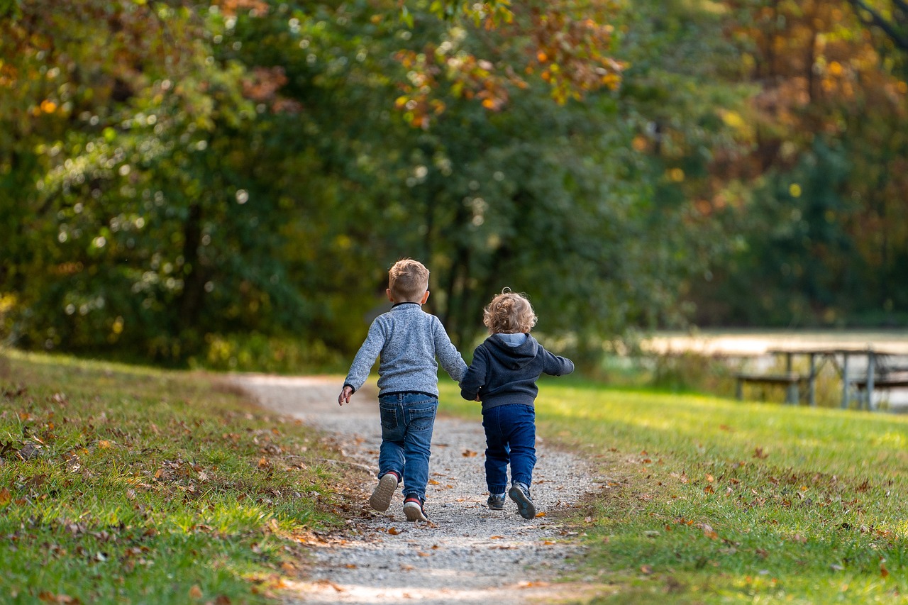 In dem Bild sind zwei kleine Kinder abgebildet, die Hand in Hand einen Kiesweg im Park entlang gehen.