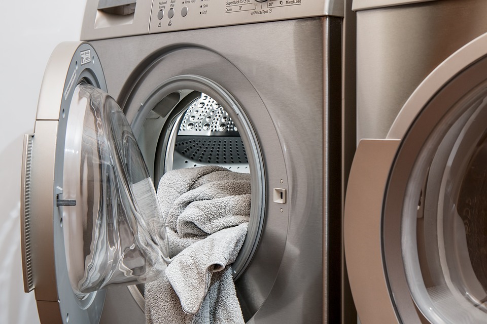 Es ist eine Waschmaschine abgebildet, deren Tür geöffnet ist und aus der Handtücher etwas heraushängen. Daneben steht ein weiteres Gerät, vermutlich ein Trockner, von dem man nur einen kleinen Ausschnitt sehen kann.