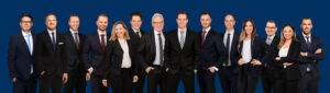 Gruppenfoto eines professionellen Teams in Businesskleidung vor blauem Hintergrund, bestehend aus sieben Männern und vier Frauen, die lächelnd und selbstbewusst in die Kamera blicken.
