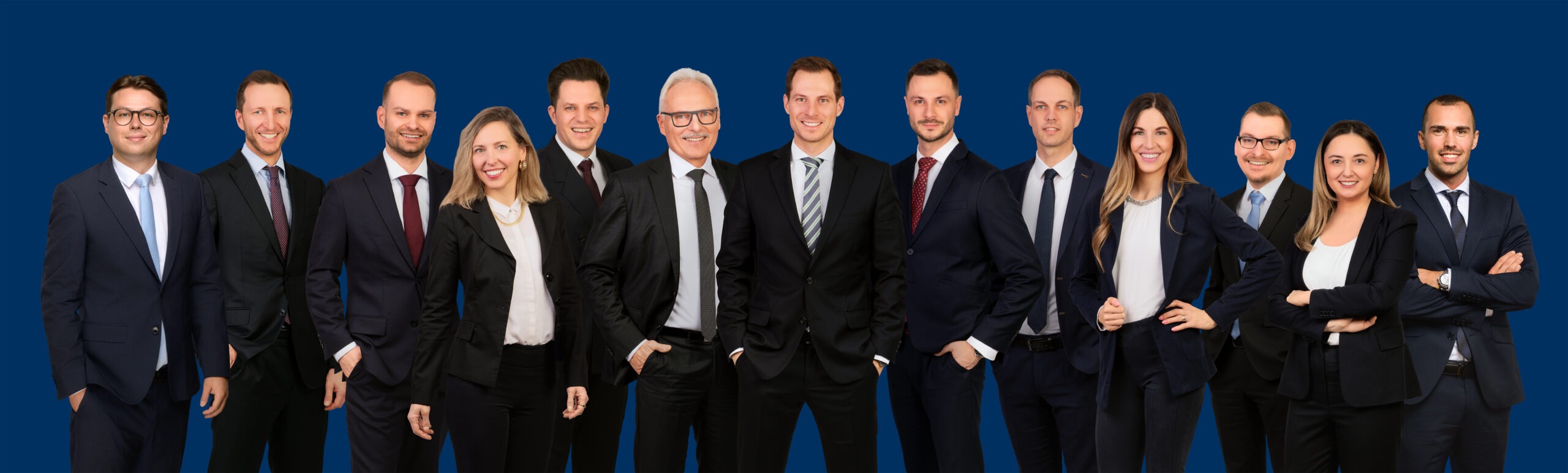 Gruppenfoto eines professionellen Teams in Businesskleidung vor blauem Hintergrund, bestehend aus sieben Männern und vier Frauen, die lächelnd und selbstbewusst in die Kamera blicken.